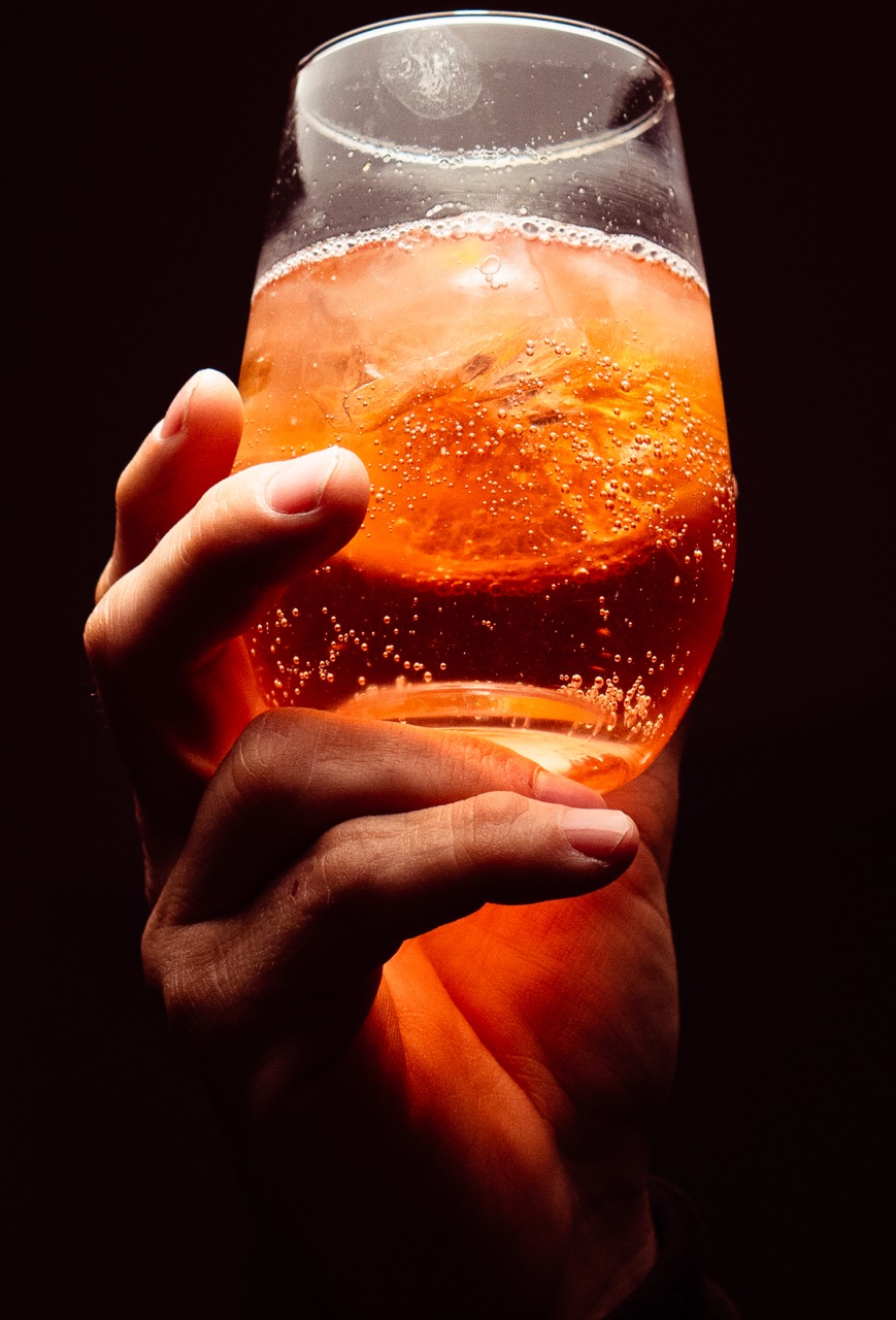 Una mano levanta un vaso de vermú con hielo y naranja a contraluz